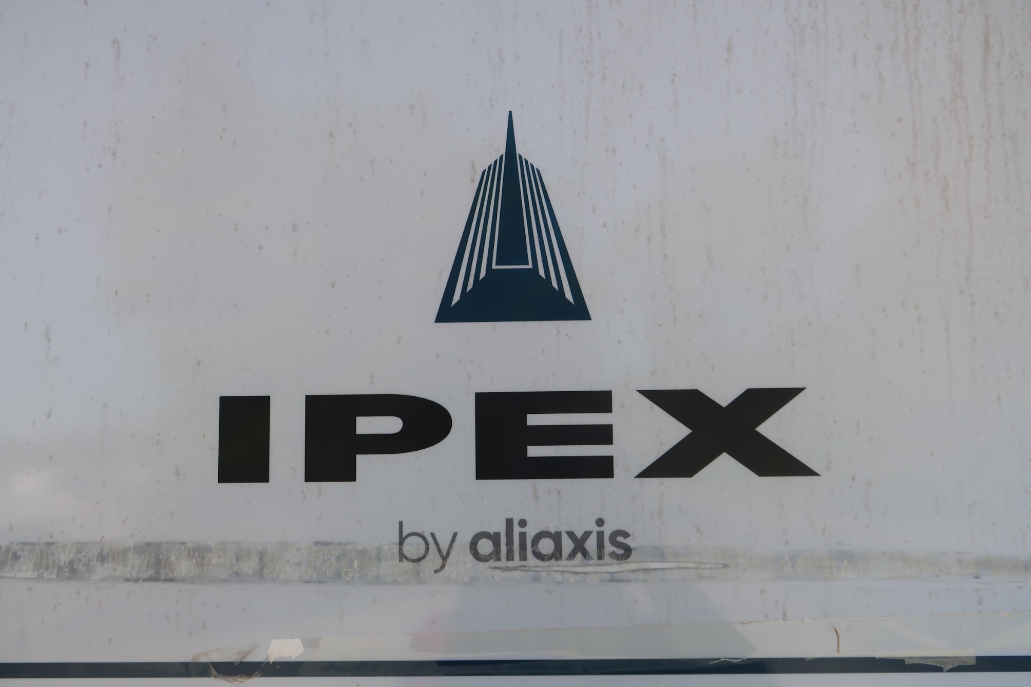 Blue triangular, pyramid-like logo with striped sides above the bold word IPEX on a weathered white sign (branding image).