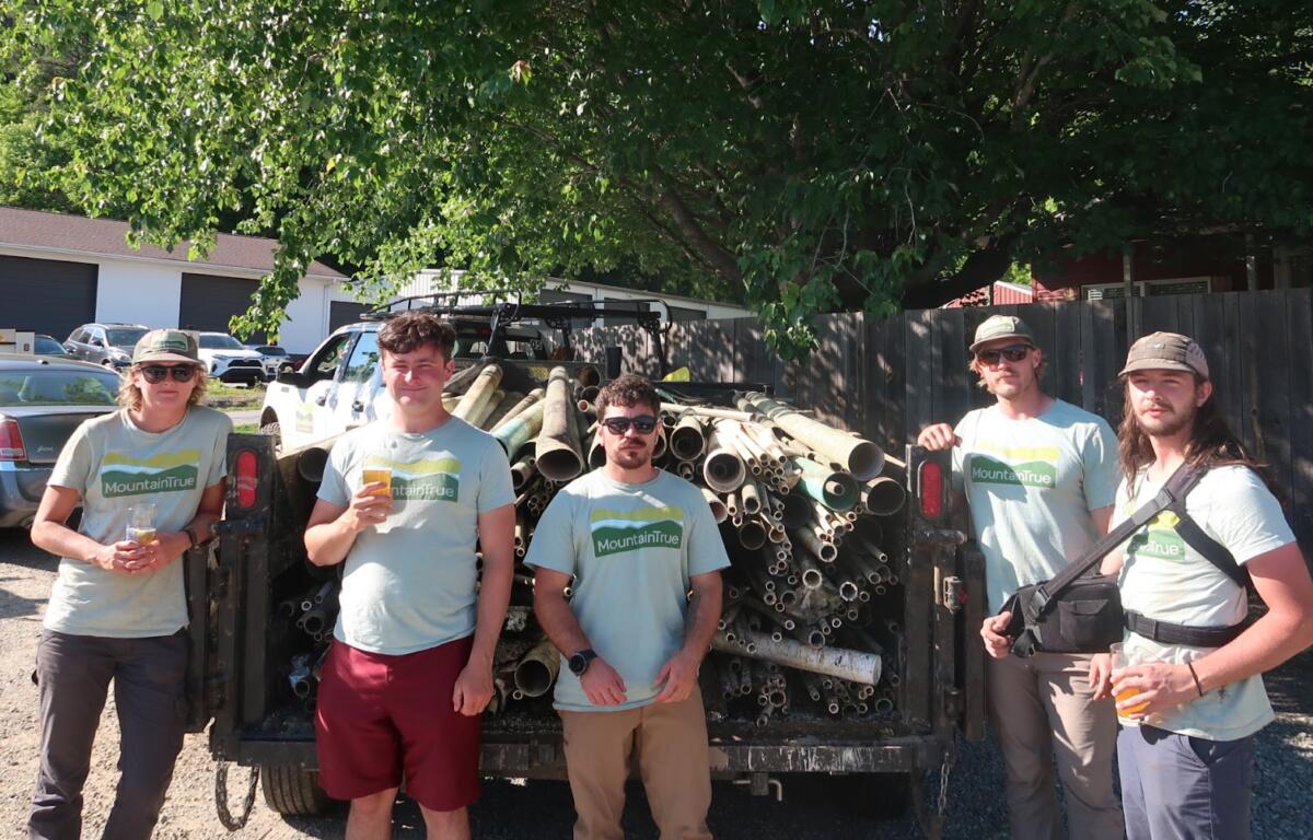 Five volunteers in Mountain True shirts stand in front of a trailer loaded with metal pipes in a sunny outdoor lot.
