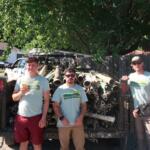 Five volunteers in Mountain True shirts stand in front of a trailer loaded with metal pipes in a sunny outdoor lot.