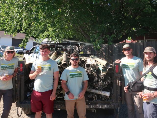 Five volunteers in Mountain True shirts stand in front of a trailer loaded with metal pipes in a sunny outdoor lot.