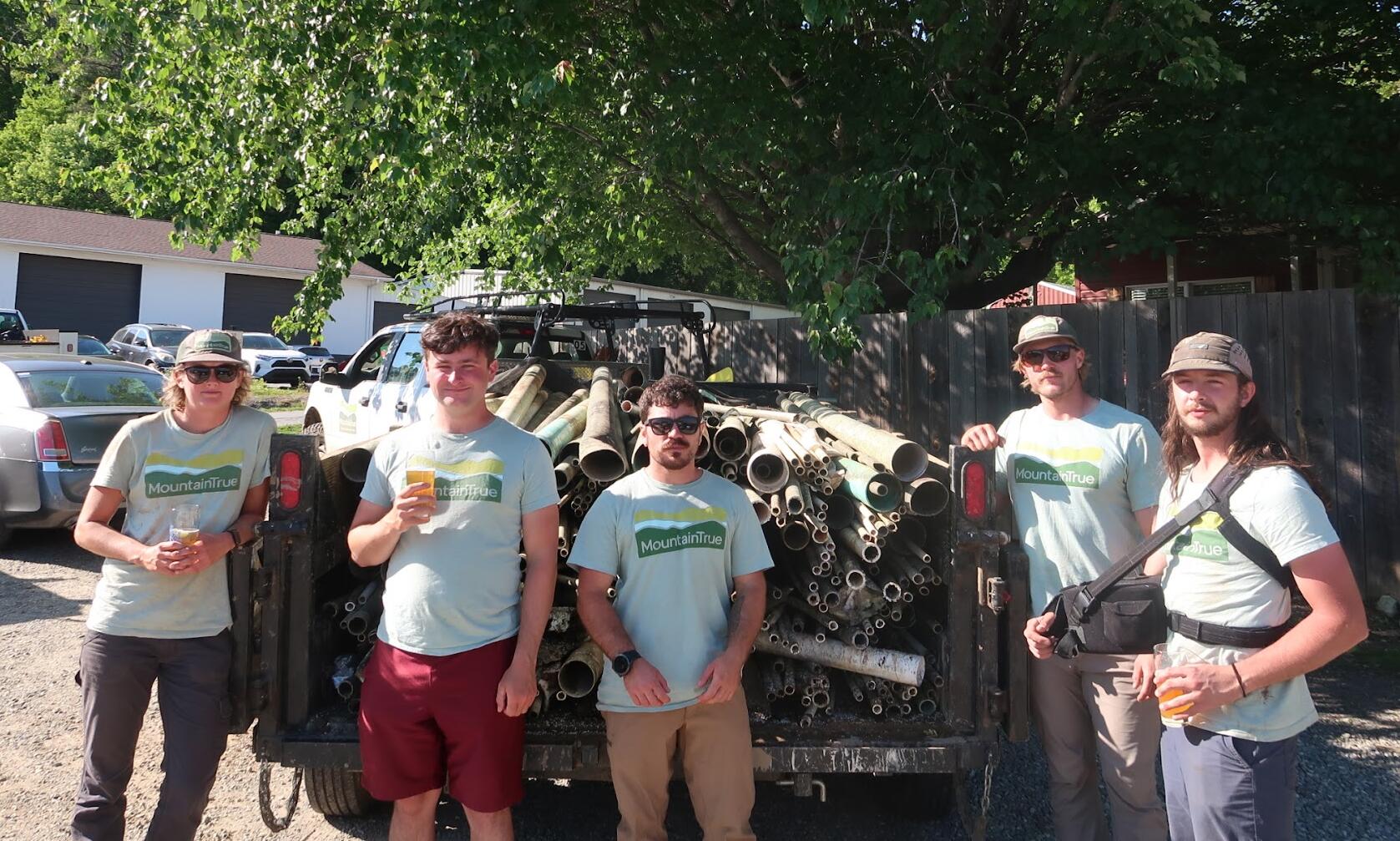 Five volunteers in Mountain True shirts stand in front of a trailer loaded with metal pipes in a sunny outdoor lot.