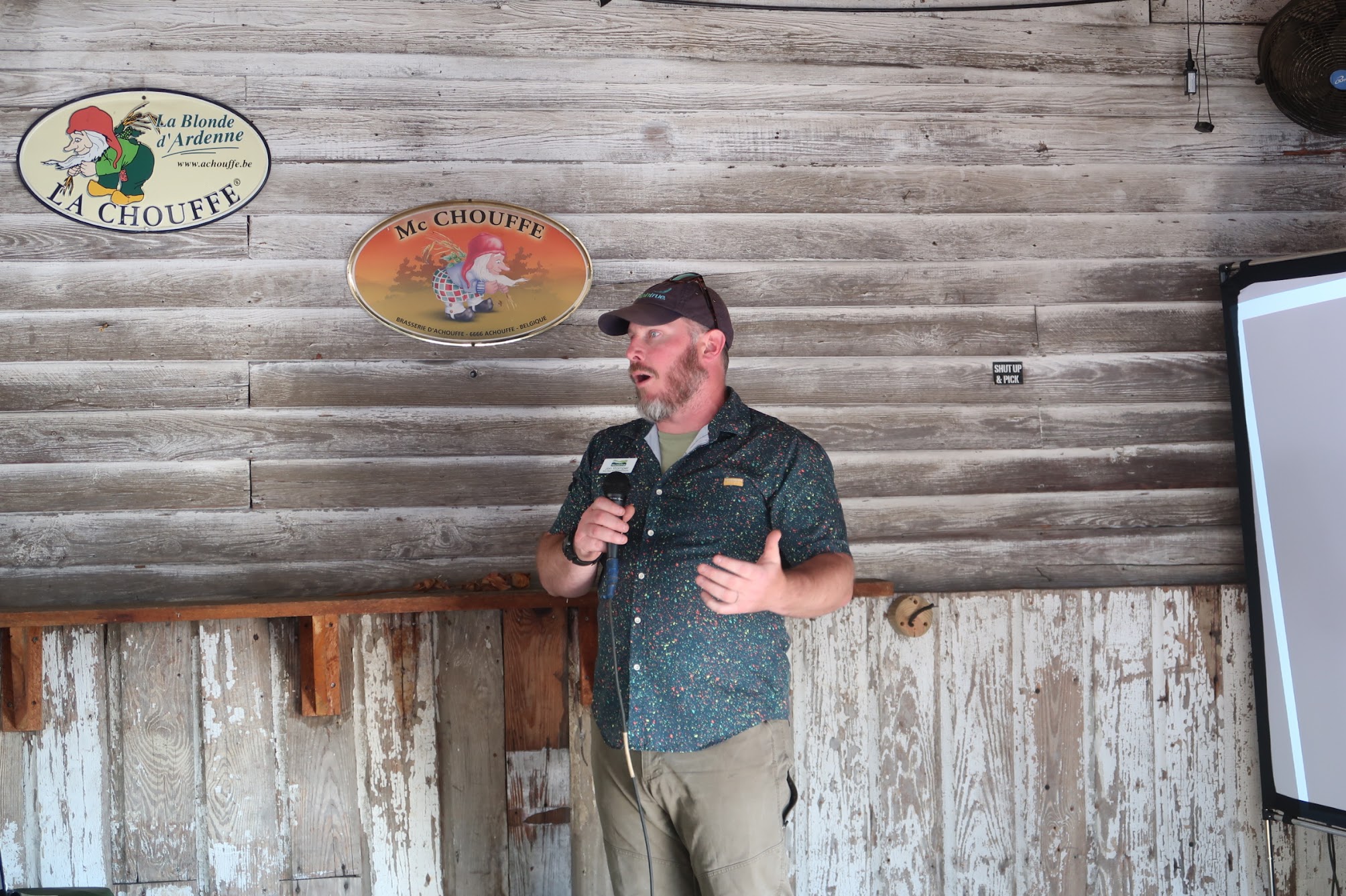 Man in a cap and patterned shirt speaks into a microphone on a wooden stage with La Chouffe and Mc Chouffe signs on the wall behind him.