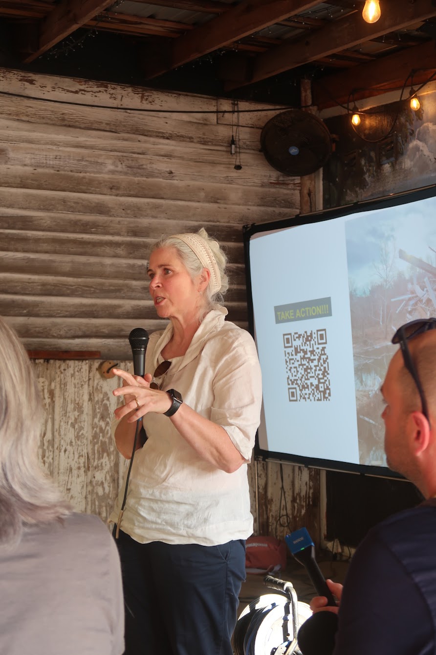 Woman in a light blouse speaks into a handheld microphone at a rustic presentation space; projector screen shows a QR code and 'TAKE ACTION!!!' text.