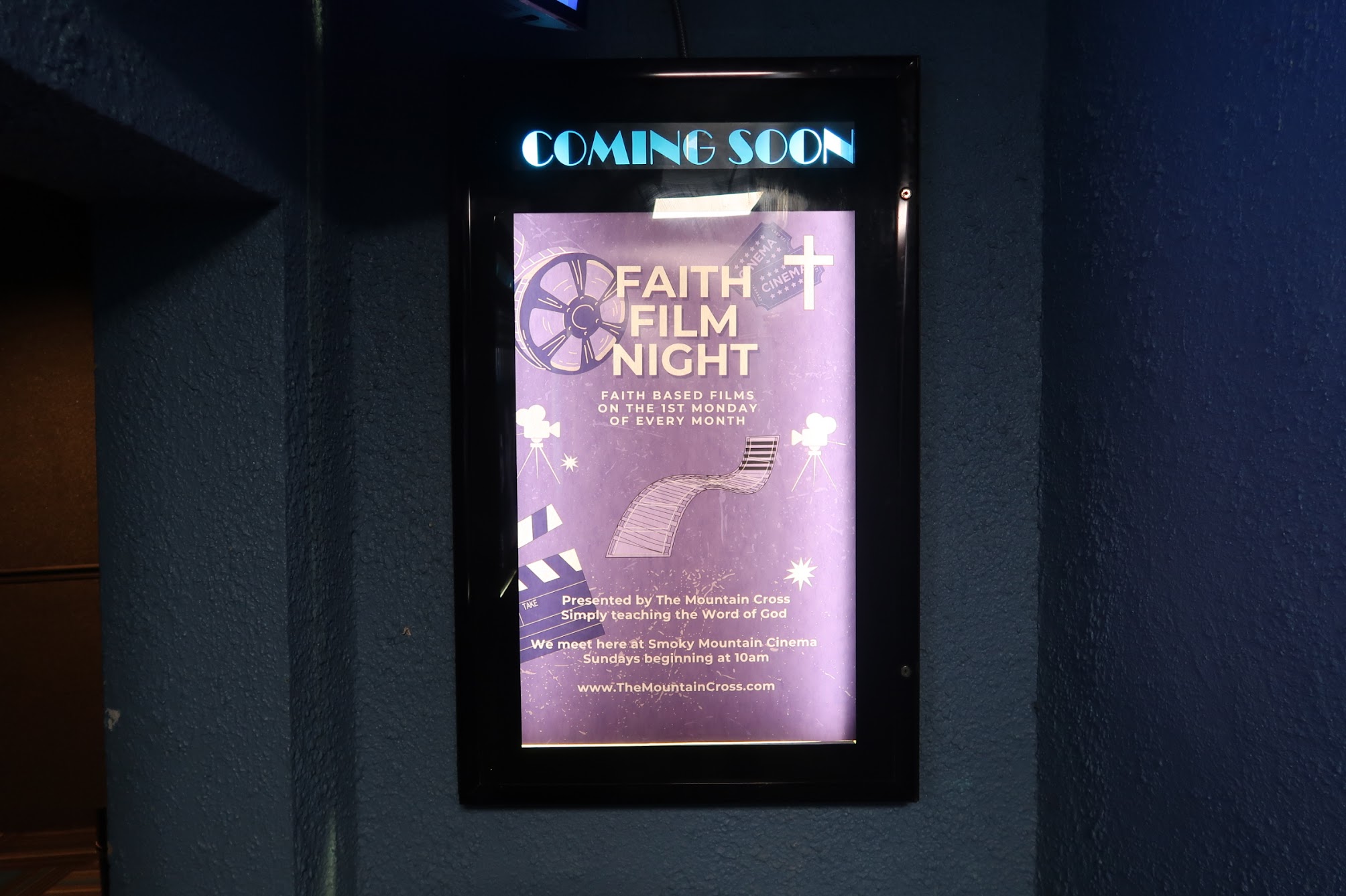 Framed purple poster on a dark blue wall advertising 'FAITH FILM NIGHT' with a cross and film reel graphics, topped by 'COMING SOON' text.