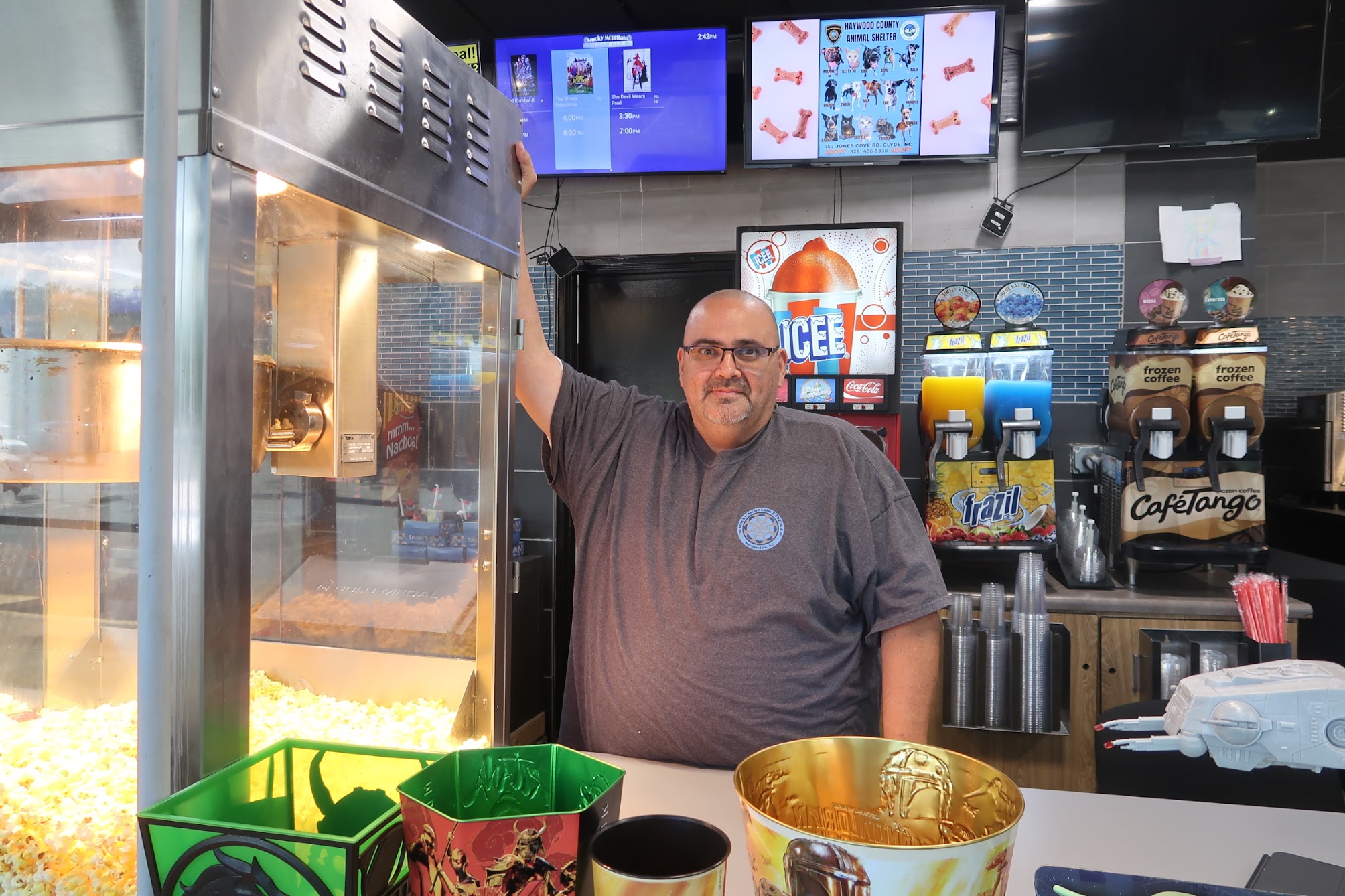 Concession stand employee behind the counter with a popcorn machine on the left and beverage dispensers to the right.