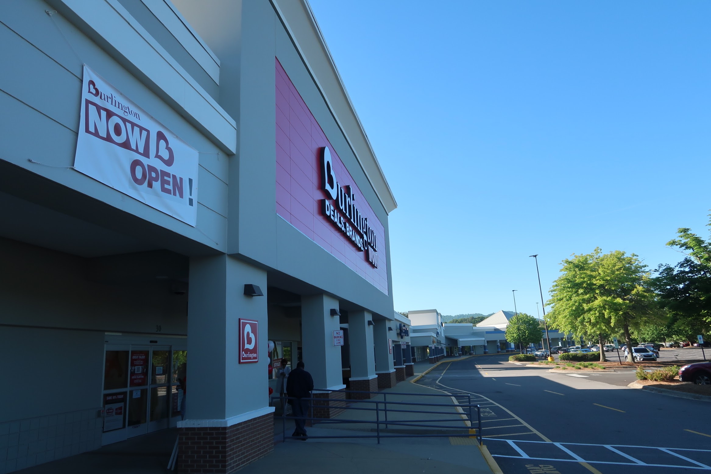 Burlington storefront with a 'NOW OPEN!' banner on the left and a purple panel façade under a clear blue sky