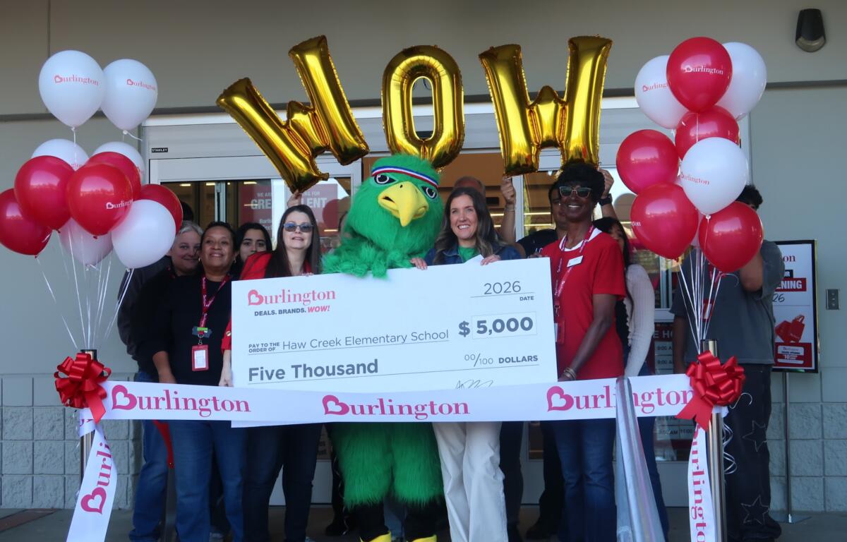 Group of people crowd around a large ceremonial check with a green mascot in the center, balloons on both sides, and Burlington branding visible all around, celebrating a donation of $5,000 to Haw Creek Elementary School.