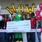Group of people crowd around a large ceremonial check with a green mascot in the center, balloons on both sides, and Burlington branding visible all around, celebrating a donation of $5,000 to Haw Creek Elementary School.