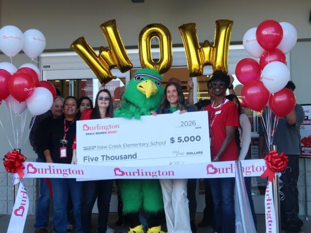 Group of people crowd around a large ceremonial check with a green mascot in the center, balloons on both sides, and Burlington branding visible all around, celebrating a donation of $5,000 to Haw Creek Elementary School.