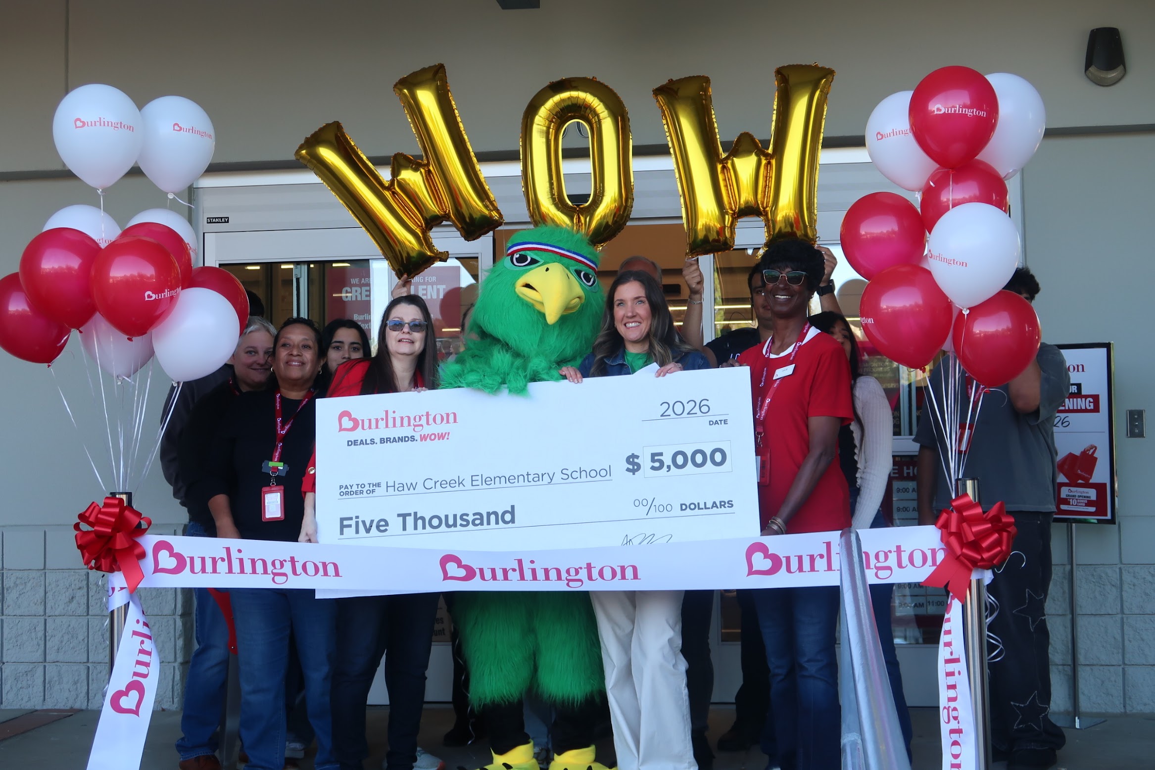 Group of people crowd around a large ceremonial check with a green mascot in the center, balloons on both sides, and Burlington branding visible all around, celebrating a donation of $5,000 to Haw Creek Elementary School.