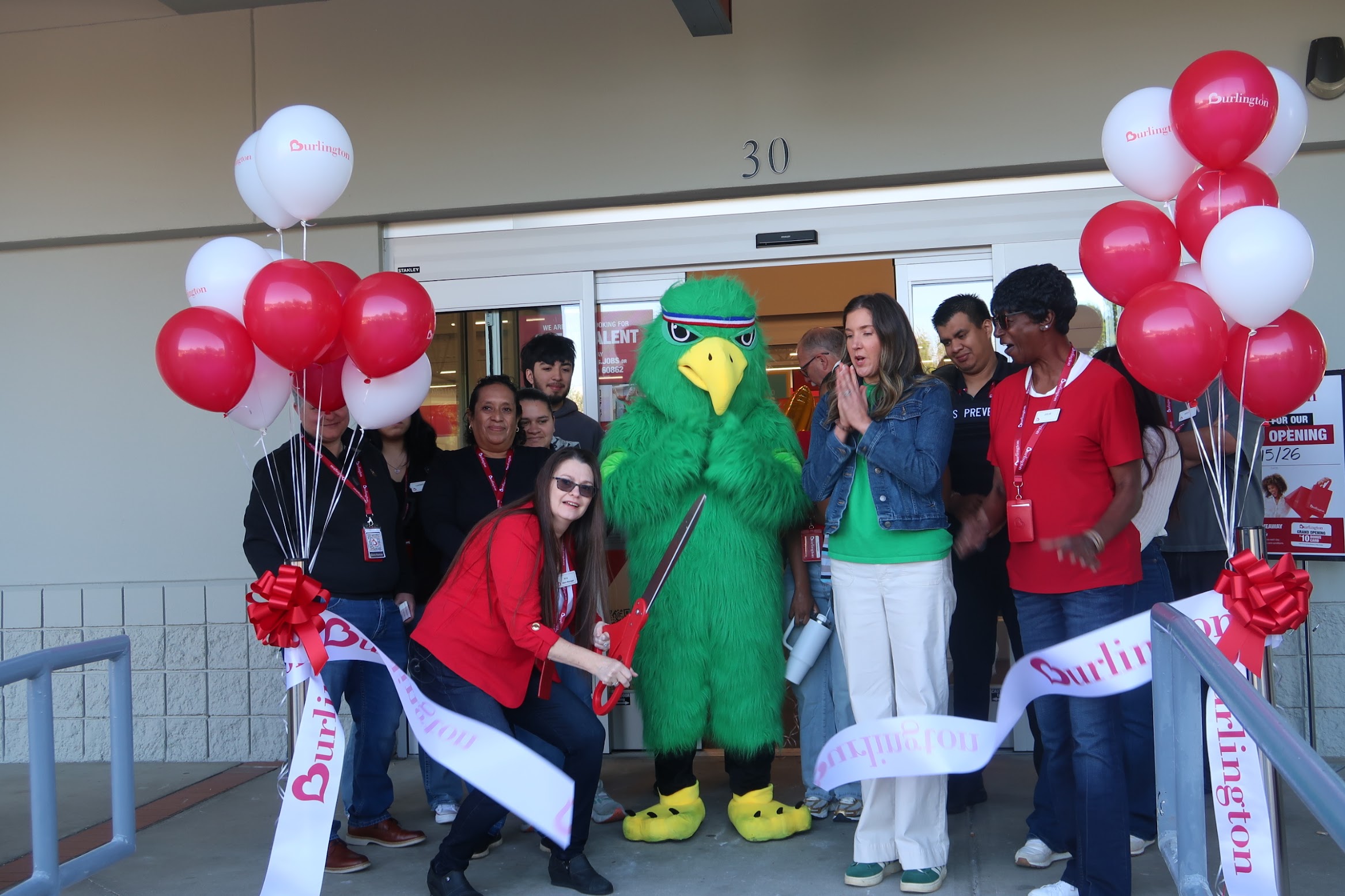Store opening celebration outside an entrance with red-and-white balloons and a green parrot mascot at the center among smiling people.