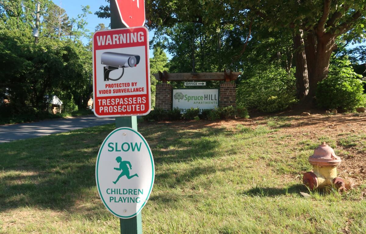 Red warning sign about video surveillance and trespassers prosecuted near the Spruce Hill Apartments entrance sign in a leafy area.