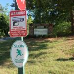 Red warning sign about video surveillance and trespassers prosecuted near the Spruce Hill Apartments entrance sign in a leafy area.