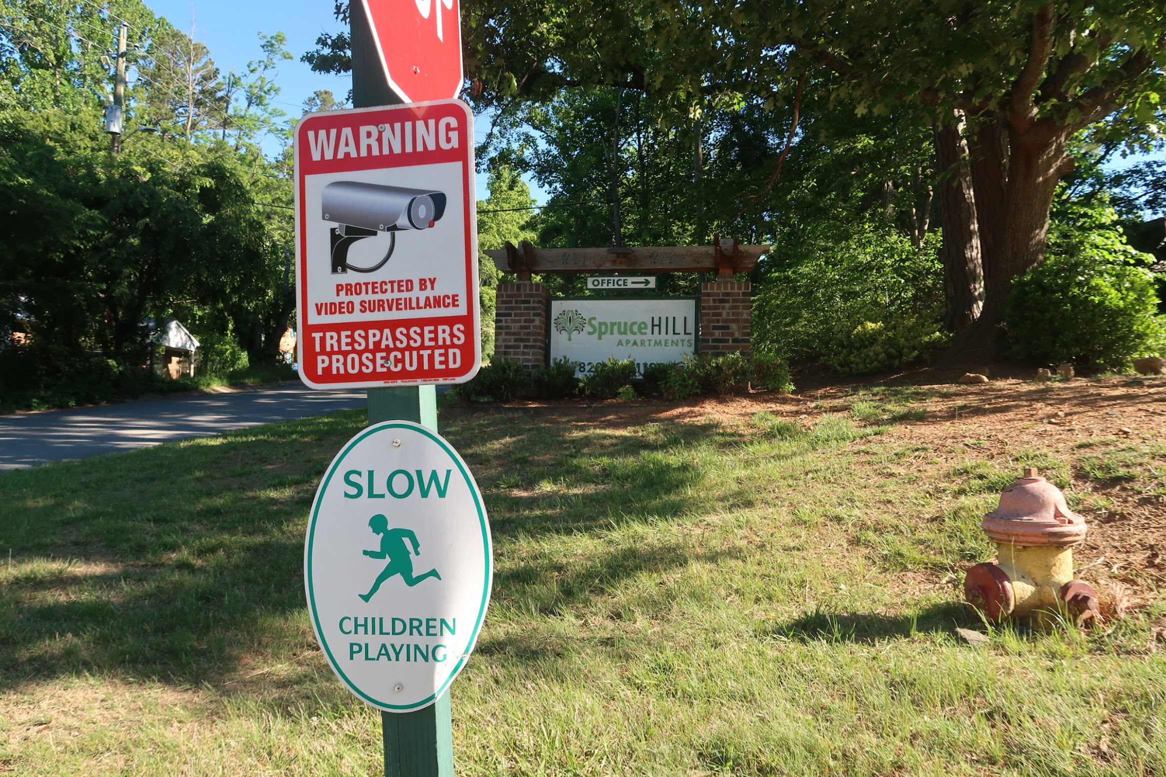 Red warning sign about video surveillance and trespassers prosecuted near the Spruce Hill Apartments entrance sign in a leafy area.