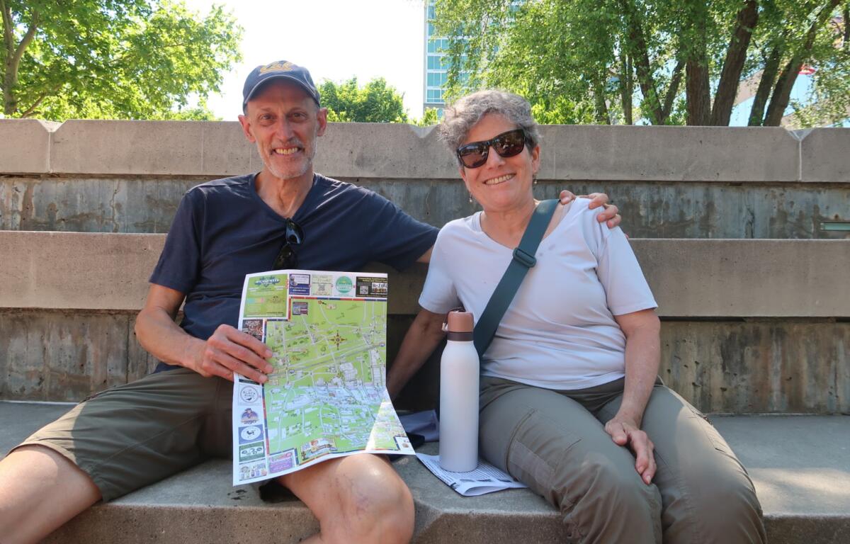A man and woman sit on outdoor concrete steps, smiling at the camera, with a colorful map and a white water bottle nearby, greenery behind them.