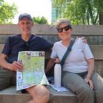 A man and woman sit on outdoor concrete steps, smiling at the camera, with a colorful map and a white water bottle nearby, greenery behind them.