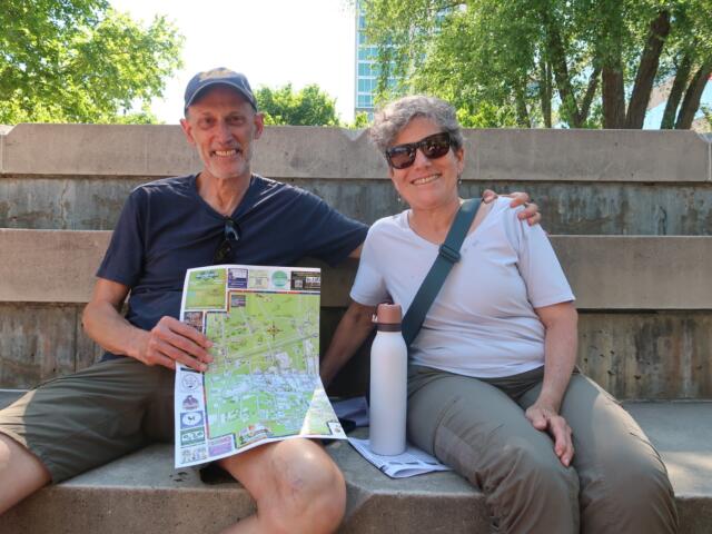 A man and woman sit on outdoor concrete steps, smiling at the camera, with a colorful map and a white water bottle nearby, greenery behind them.