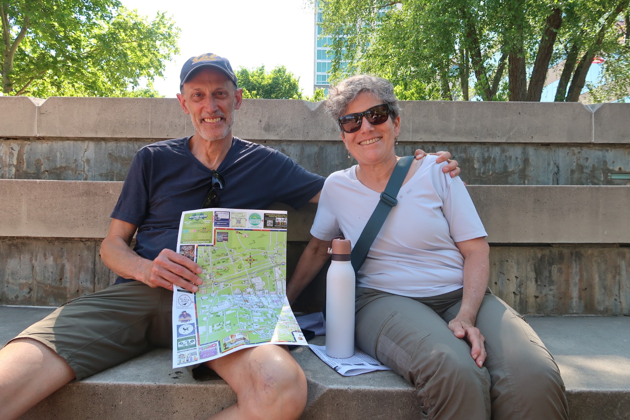 A man and woman sit on outdoor concrete steps, smiling at the camera, with a colorful map and a white water bottle nearby, greenery behind them.