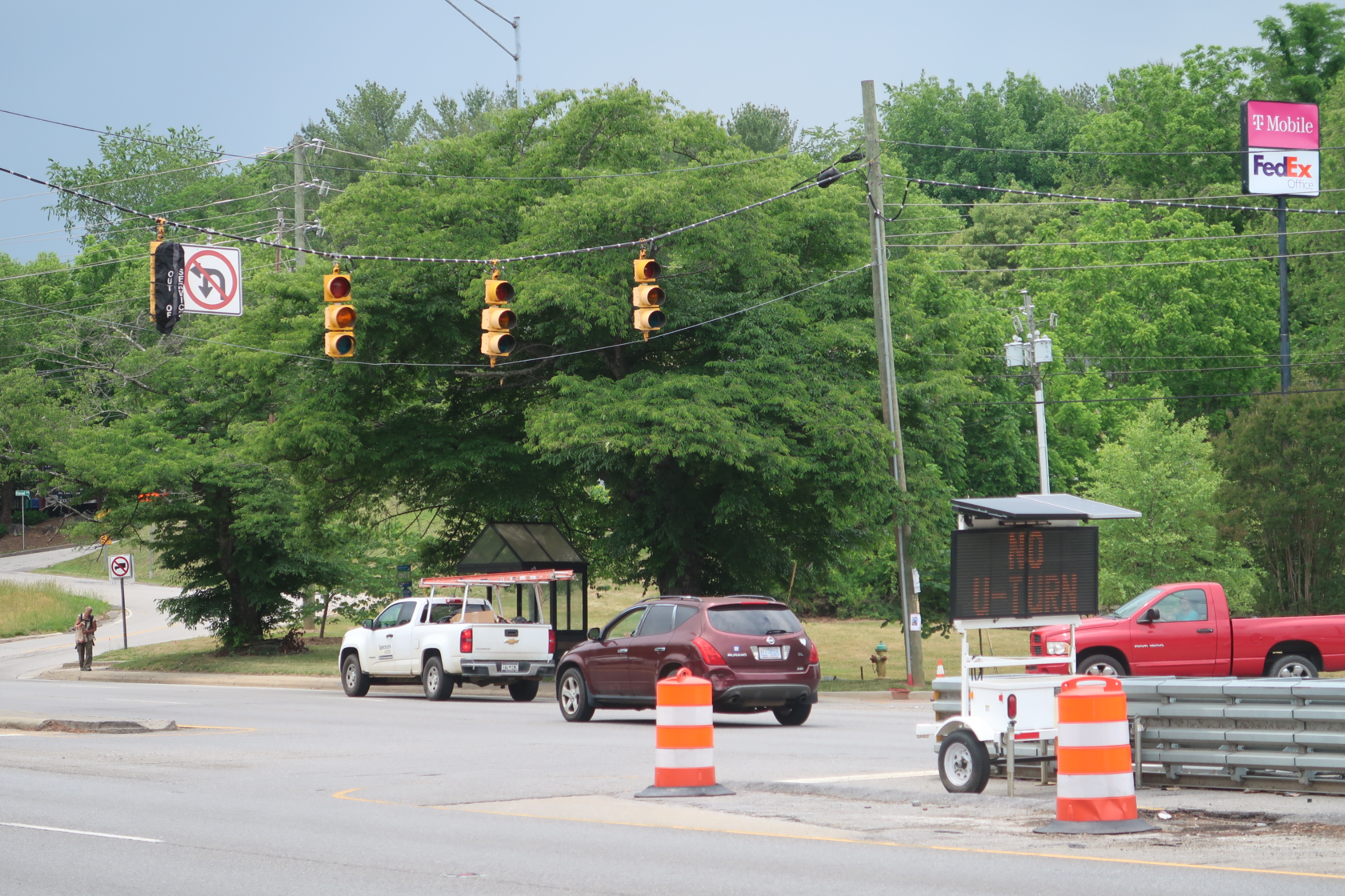 Urban intersection with orange construction barrels, traffic lights, and a digital display sign among green trees.