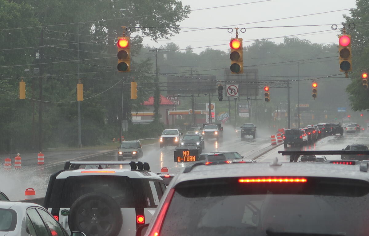 Rainy city intersection with multiple red lights, cars in traffic, and orange construction cones on the wet road.