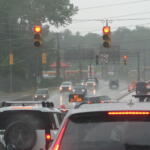 Rainy city intersection with multiple red lights, cars in traffic, and orange construction cones on the wet road.