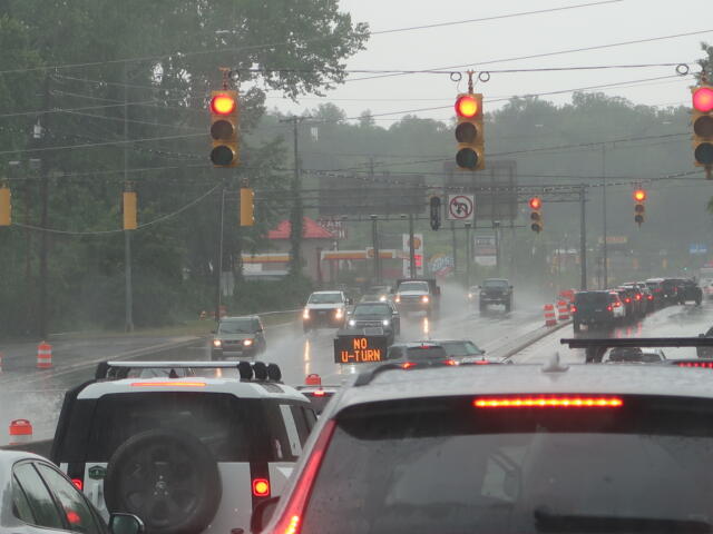 Rainy city intersection with multiple red lights, cars in traffic, and orange construction cones on the wet road.
