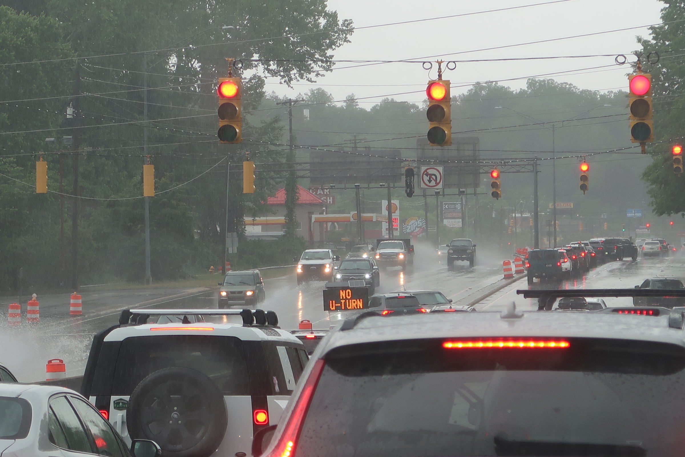 Rainy city intersection with multiple red lights, cars in traffic, and orange construction cones on the wet road.