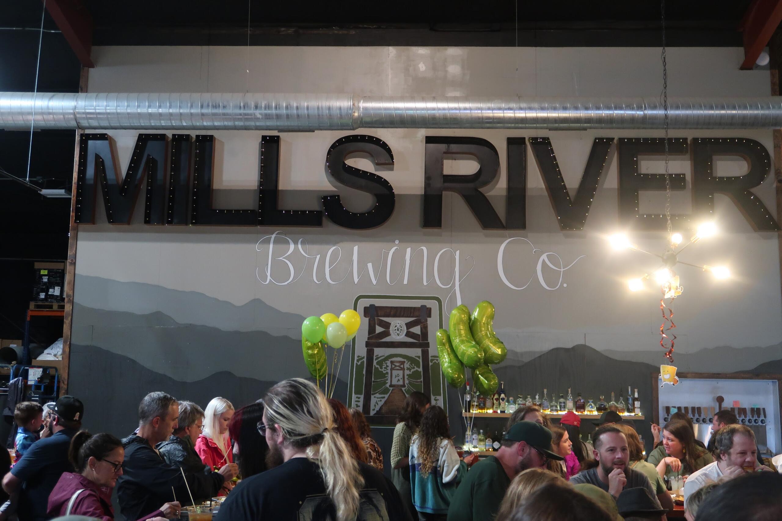 Crowded brewery event beneath a large sign reading 'Miles River Brewing Co.' with balloons and a busy bar area behind the crowd.