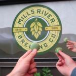 Hands holding several green macarons in front of a circular Mills River Brewing Co. logo on a window.