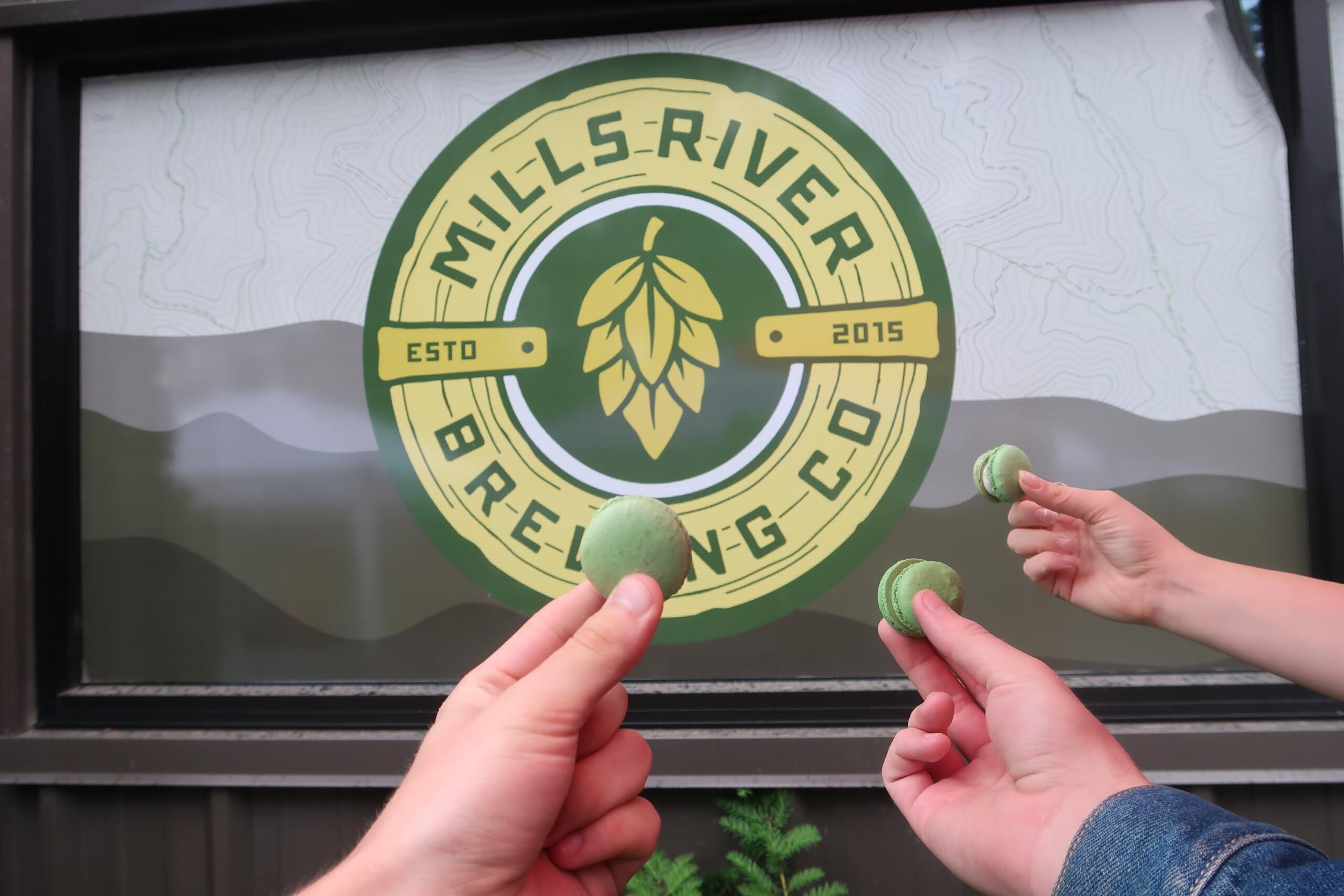Hands holding several green macarons in front of a circular Mills River Brewing Co. logo on a window.