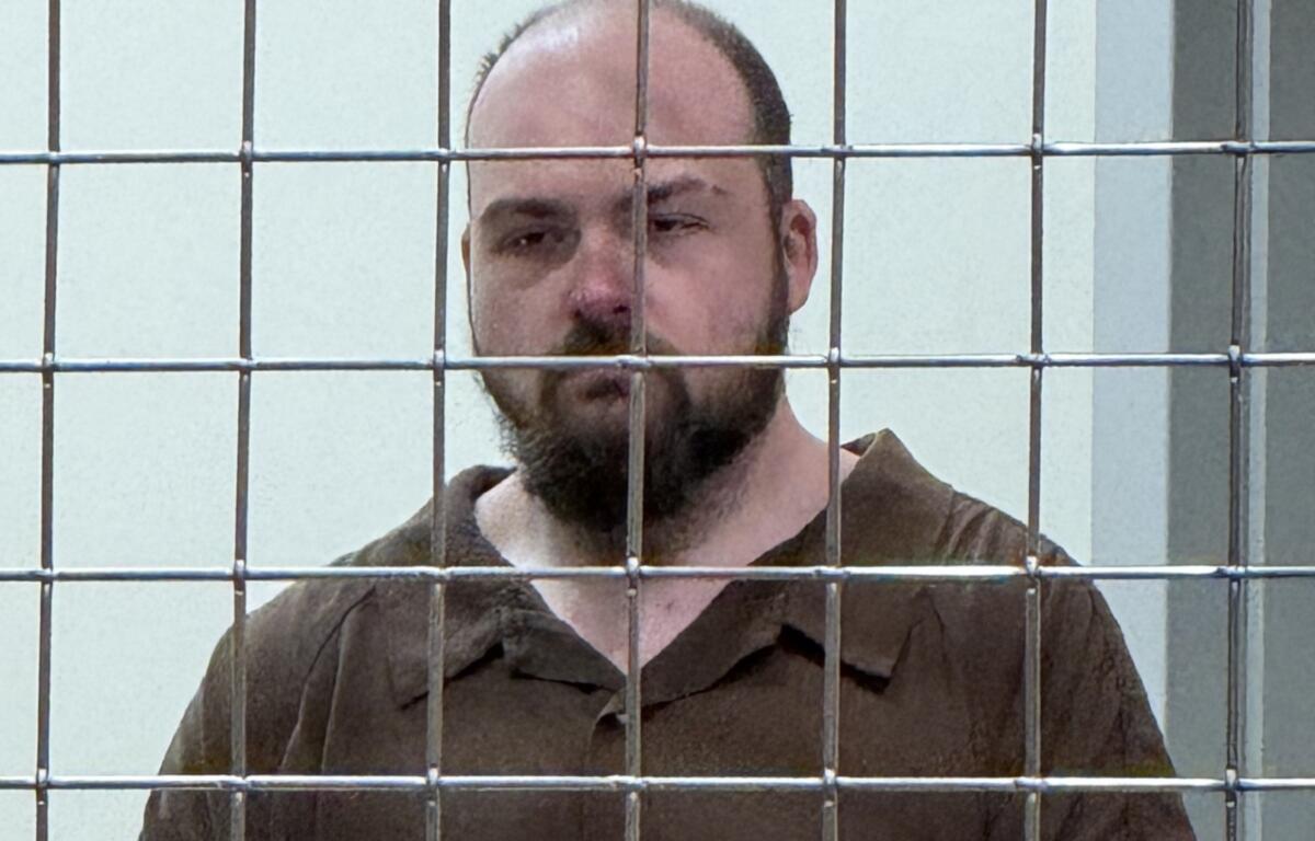 Bearded man in a brown prison jumpsuit behind a metal bar enclosure, looking toward the camera.