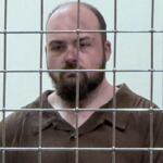 Bearded man in a brown prison jumpsuit behind a metal bar enclosure, looking toward the camera.