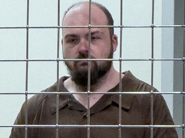 Bearded man in a brown prison jumpsuit behind a metal bar enclosure, looking toward the camera.
