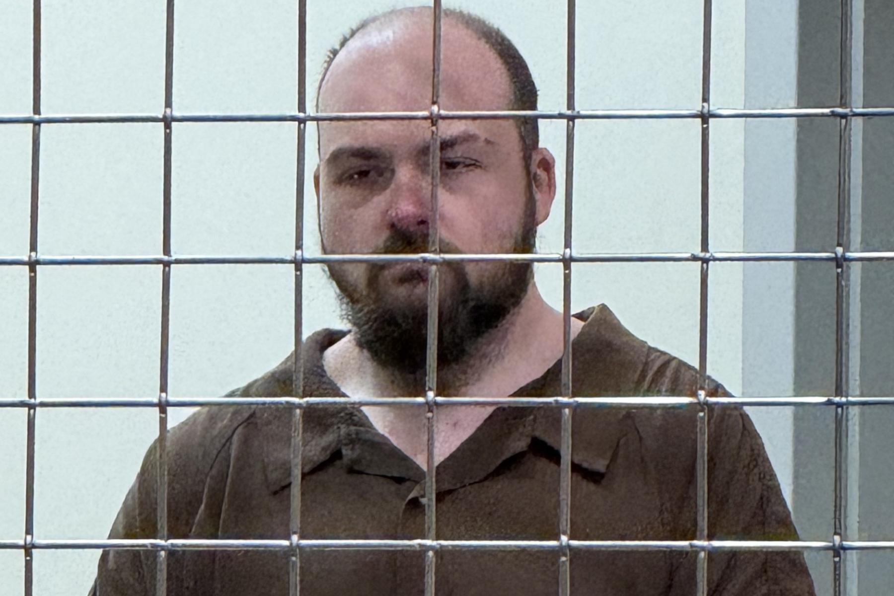 Bearded man in a brown prison jumpsuit behind a metal bar enclosure, looking toward the camera.