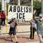 Group of protesters on steps holding a large 'ABOLISH ICE' banner and a sign reading 'Is it fascism yet?'.