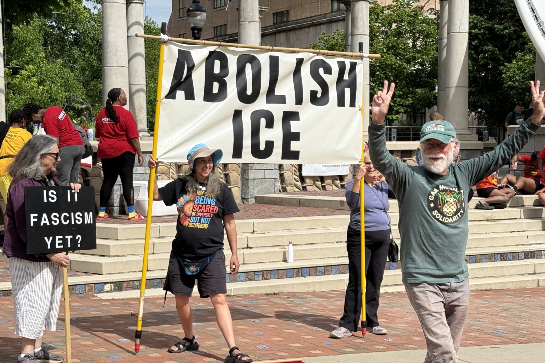 Group of protesters on steps holding a large 'ABOLISH ICE' banner and a sign reading 'Is it fascism yet?'.