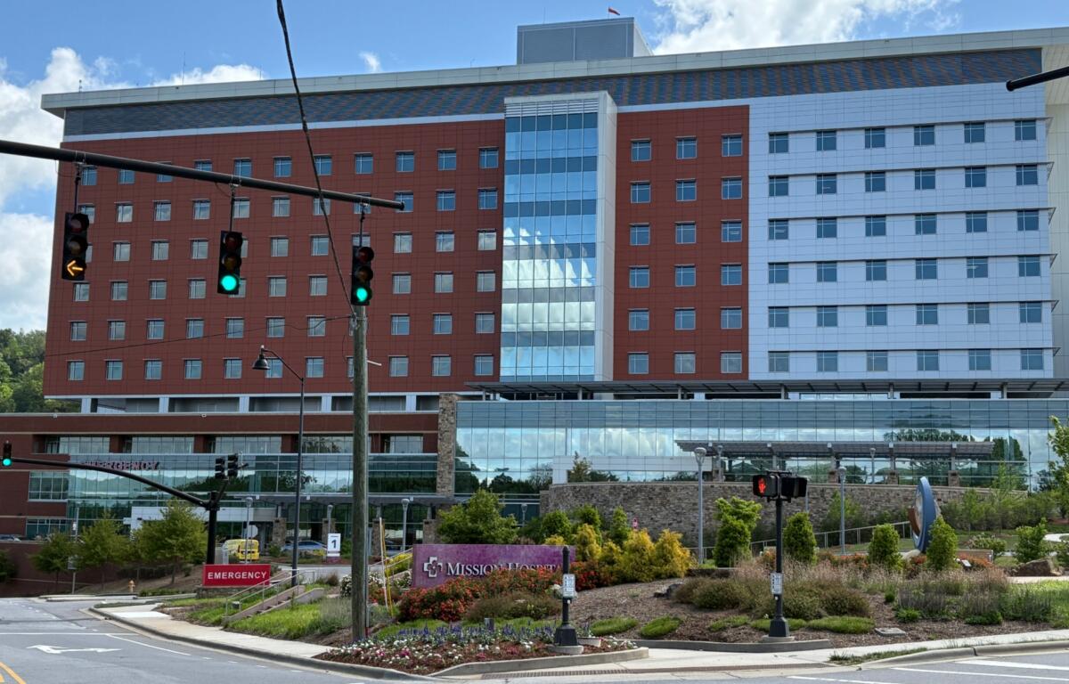 Modern hospital building with red brick wings, blue glass atrium, and an emergency entrance facing the street with blue sky above