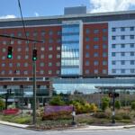 Modern hospital building with red brick wings, blue glass atrium, and an emergency entrance facing the street with blue sky above