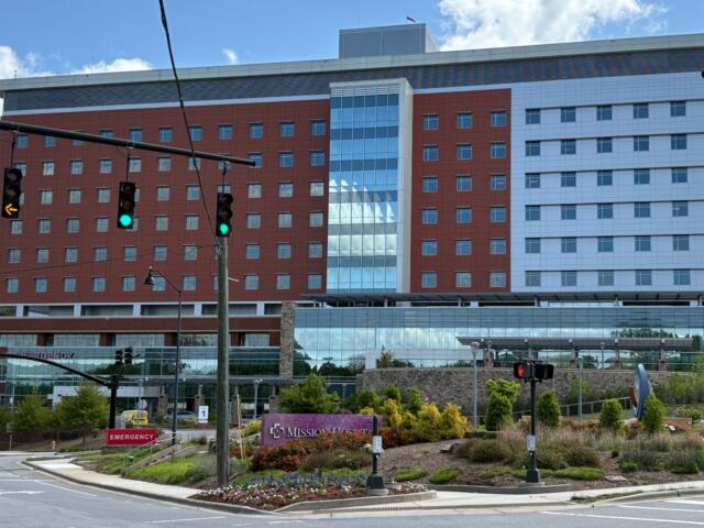 Modern hospital building with red brick wings, blue glass atrium, and an emergency entrance facing the street with blue sky above