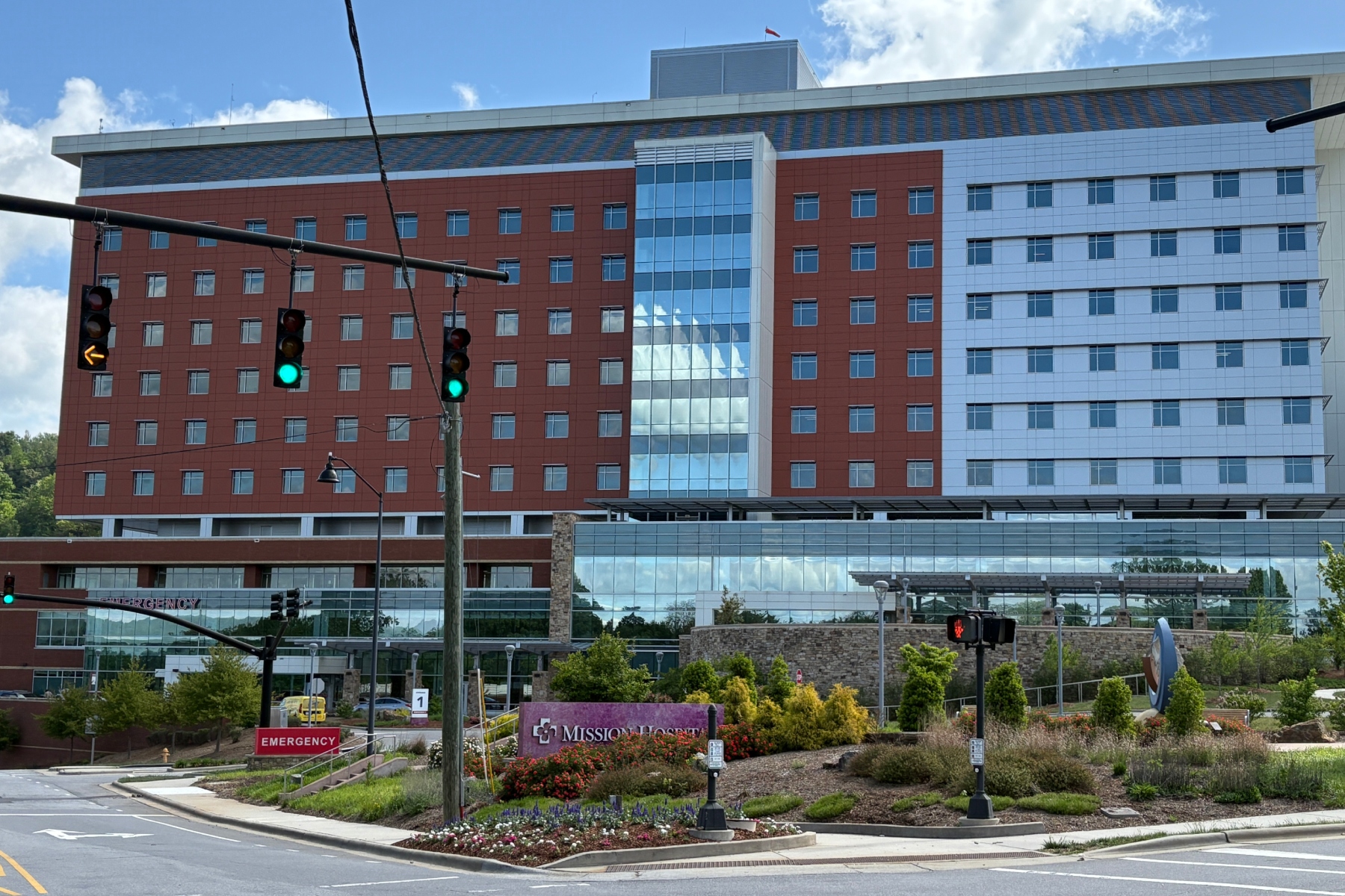 Modern hospital building with red brick wings, blue glass atrium, and an emergency entrance facing the street with blue sky above