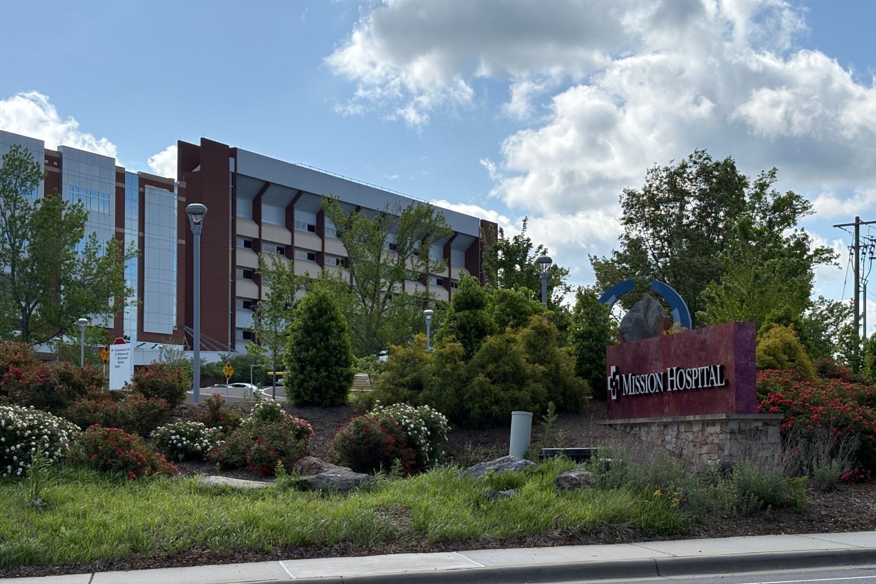 Mission Hospital sign in front of landscaped gardens and a modern brick building under a blue sky with clouds, a welcoming campus view.