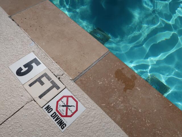 Pool deck with warning signs near the water edge, including 'No diving' and 'Too shallow' messages.