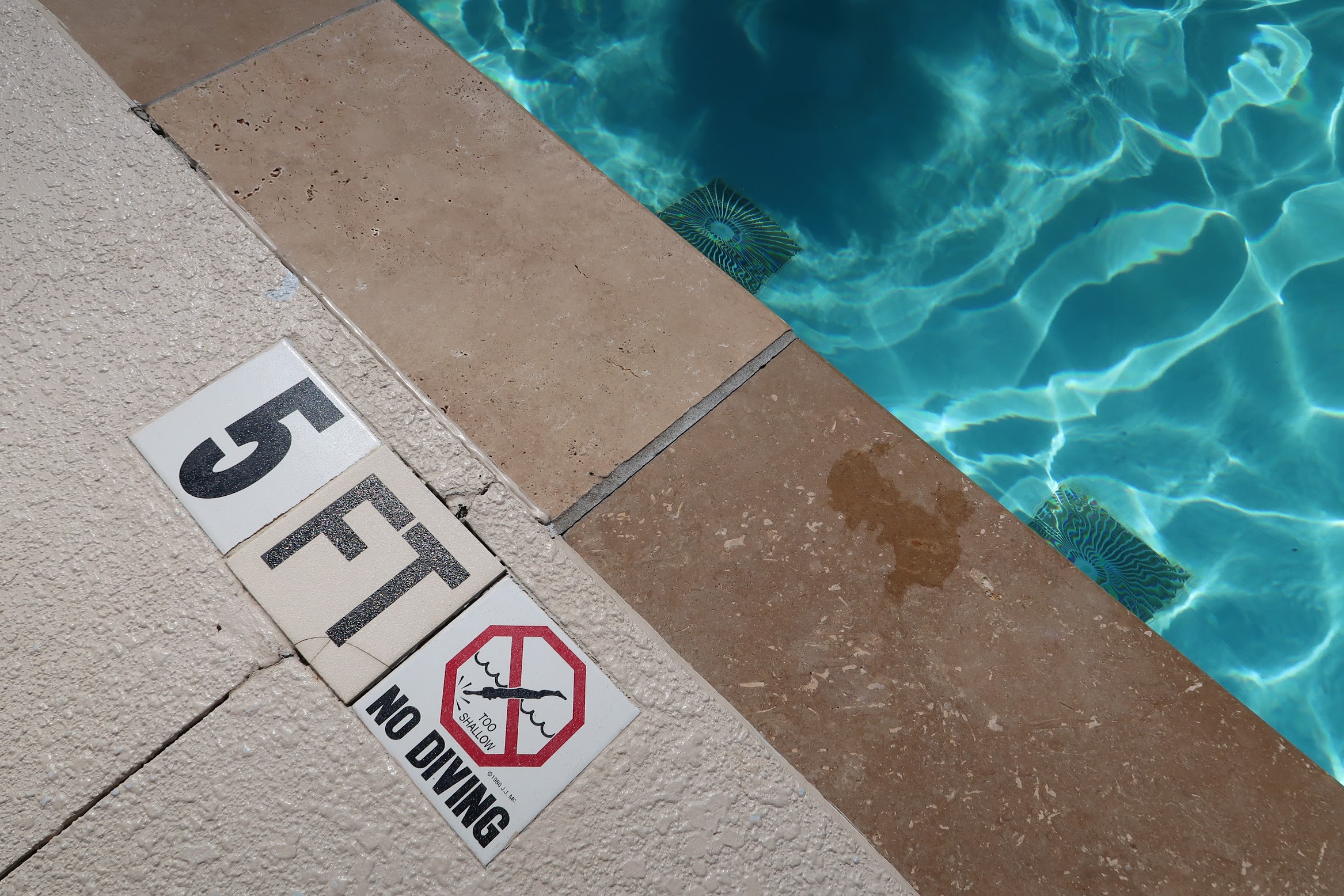 Pool deck with warning signs near the water edge, including 'No diving' and 'Too shallow' messages.