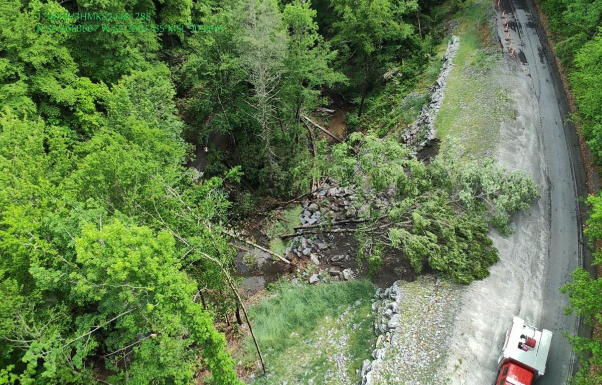 Aerial view of a forested hillside with rock rubble along a winding road and a red emergency vehicle at the bottom right.