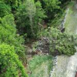 Aerial view of a forested hillside with rock rubble along a winding road and a red emergency vehicle at the bottom right.