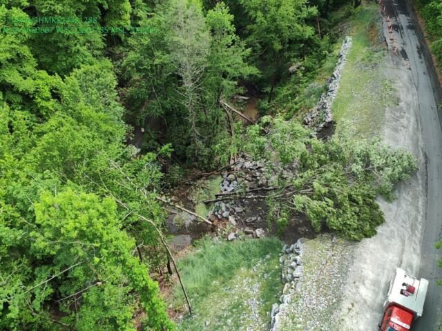 Aerial view of a forested hillside with rock rubble along a winding road and a red emergency vehicle at the bottom right.