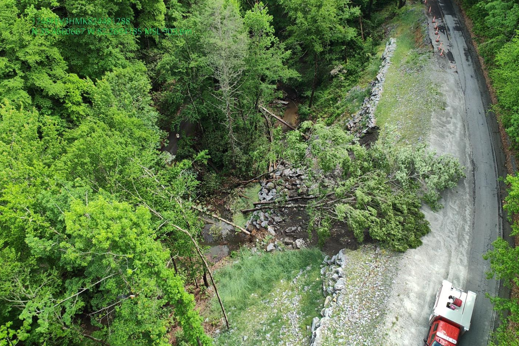 Aerial view of a forested hillside with rock rubble along a winding road and a red emergency vehicle at the bottom right.