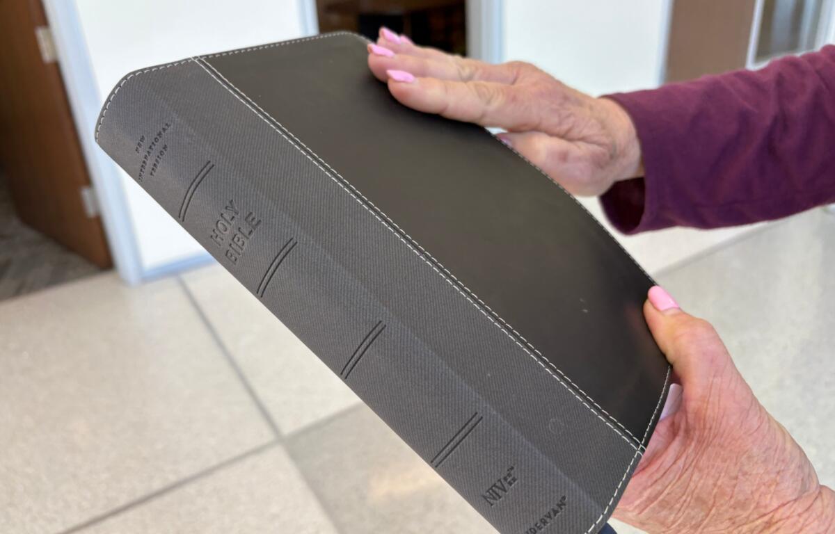 A person with pink nails and a purple sleeve holding a black leather Bible with white stitching and embossed title.