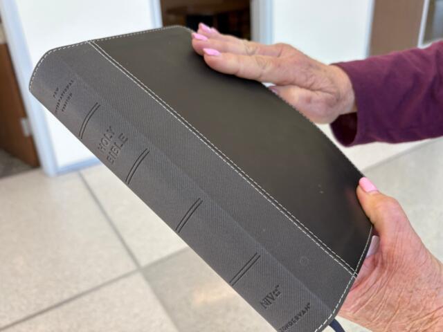 A person with pink nails and a purple sleeve holding a black leather Bible with white stitching and embossed title.