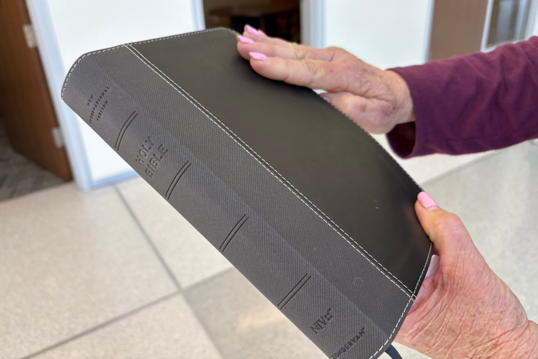 A person with pink nails and a purple sleeve holding a black leather Bible with white stitching and embossed title.